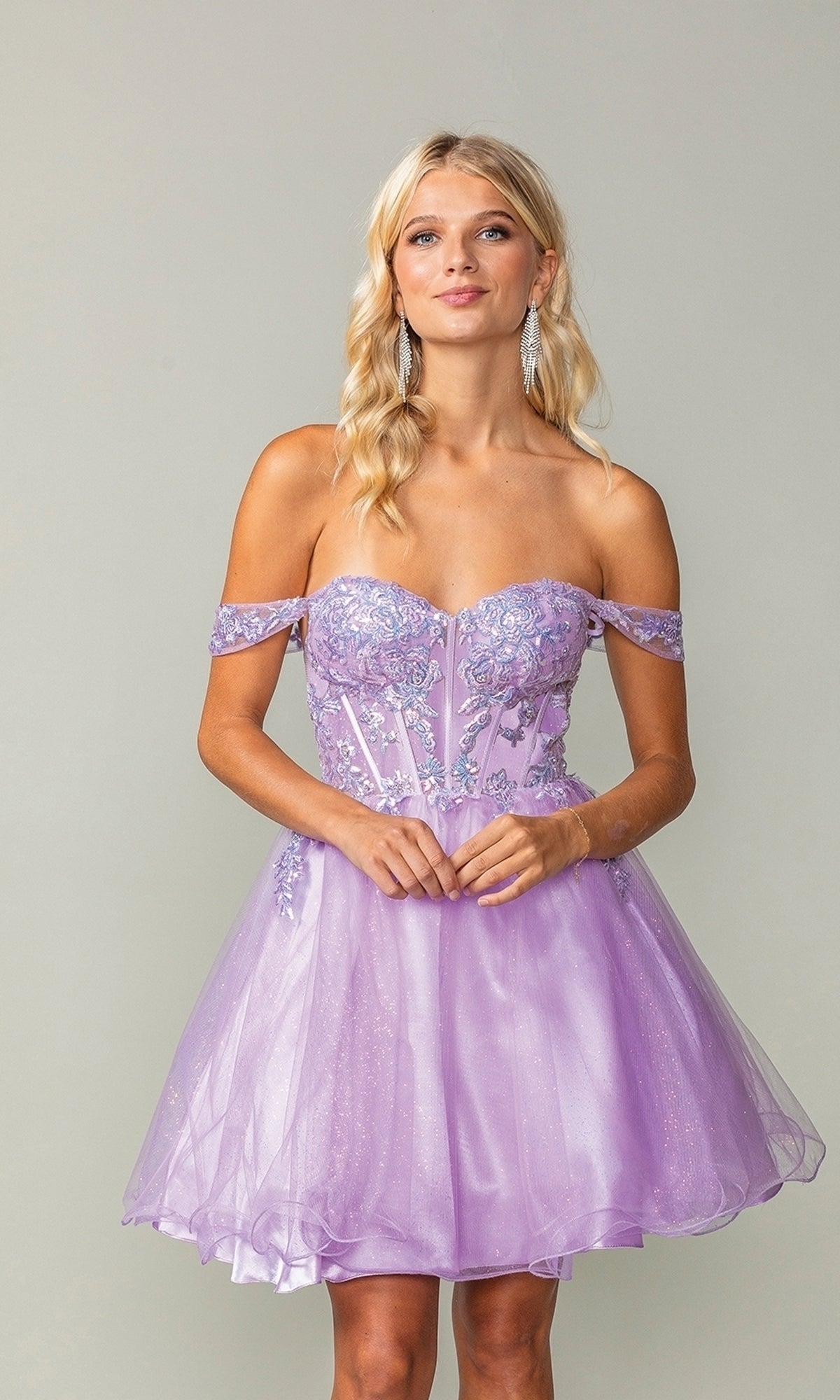A woman wears the Dancing Queen 3383 Short Corset Homecoming Dress—an off-shoulder lavender dress with floral embroidery and a sparkly sheer corset bodice—standing against a plain light background.
